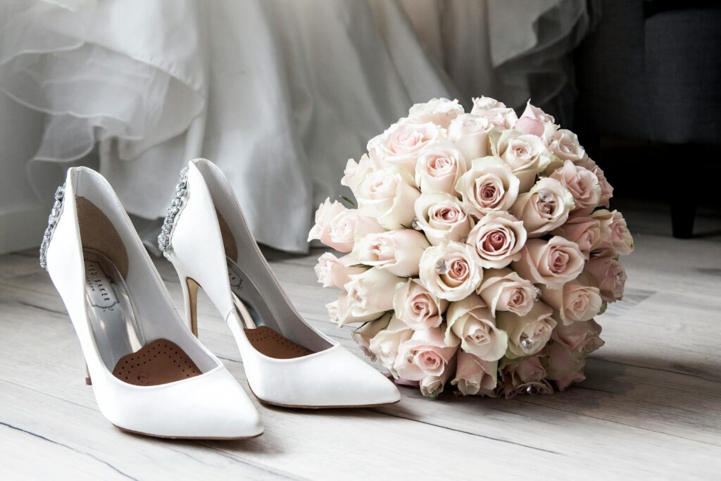 Ein weißes Paar Pumps mit einem Strauß Rosen nebenbei, im Hintergrund sieht man das untere ende eines Kleides
